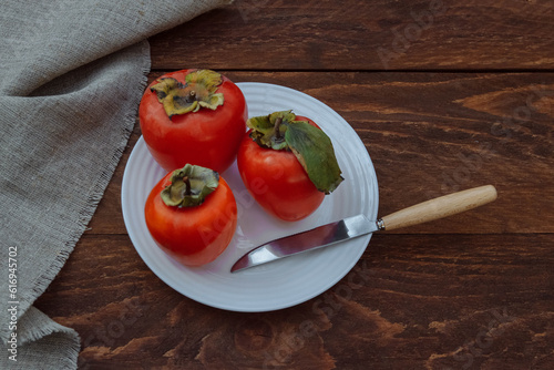 Ripe, juicy and red persimmon with green leaves on a white plate with a knife and linen cloth on a brown wooden background, seasonal fruit concept, vitamins, autumn harvest, healthy food
