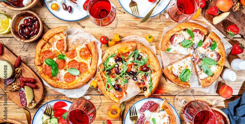 Pizza party table. Top view glasses with red wine and dinner plates on rustic wooden table with hot pizzas, italian appetizers, salads, cheese, fruits and berries. Family lunch with fast food. Banner