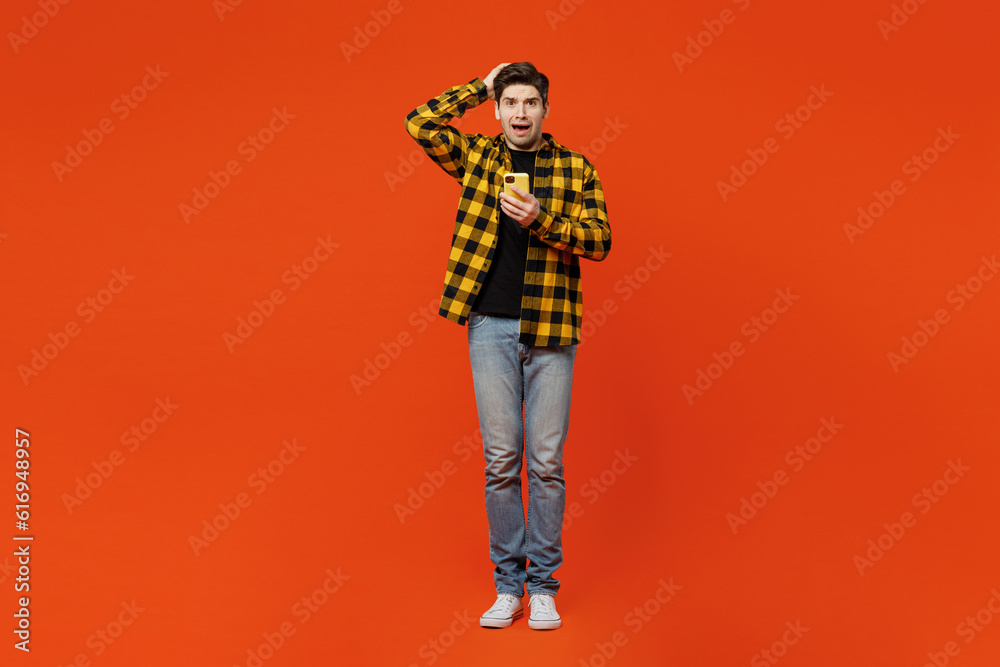 Full body scared sad uhappy young caucasian man he wear yellow checkered shirt black t-shirt hold head use mobile cell phone isolated on plain red orange background studio portrait. Lifestyle concept. Full body scared sad uhappy young caucasian man he wear yellow checkered shirt black t-shirt hold head use mobile cell phone isolated on plain red orange background studio portrait. Lifestyle concept.
