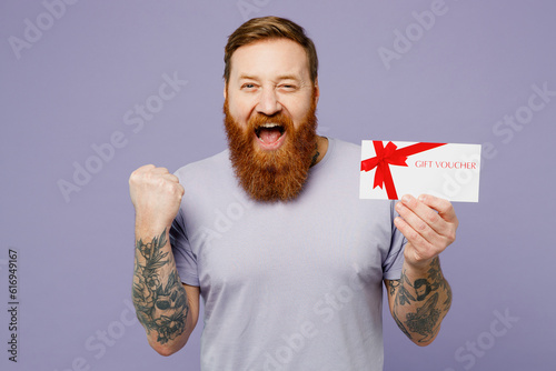 Young redhead bearded man wear violet t-shirt casual clothes hold gift certificate coupon voucher card for store do winner gesture isolated on plain pastel light purple background Lifestyle concept.