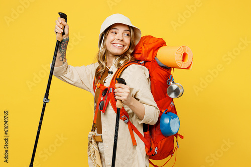 Young woman carry backpack with stuff mat holding trekking poles look aside isolated on plain yellow background Tourist leads active lifestyle walk on spare time. Hiking trek rest travel trip concept #616949765