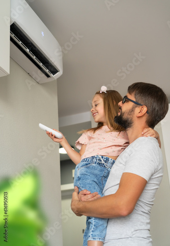 Happy little girl holds remote control from air conditioner sitting in fathers hands in light interior of apartment. Family turn on air conditioner