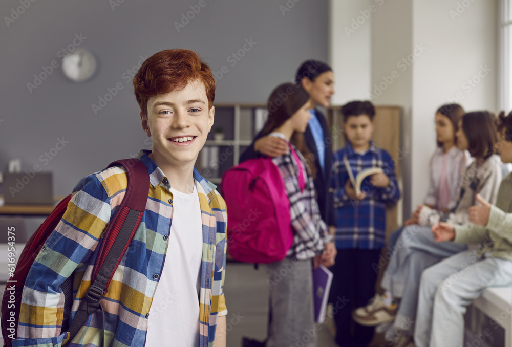 Fototapeta premium Portrait of smiling redhead boy standing with backpack in classroom. Happy schoolboy wearing casual plaid shirt posing on background of his classmates and teacher and looking at camera