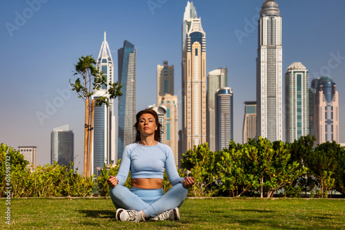 Middle age sporty woman sitting lotus yoga pose on lawn in urban park at Dubai skyscrapers. Cute lady training in city environment, outdoor workout. Healthy lifestyle concept. Copy ad text space © Alex Vog
