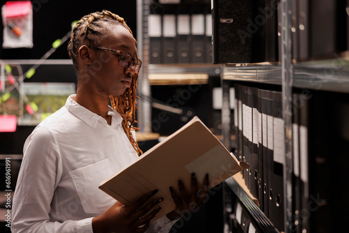 Fotografi African american woman law enforcement professional reading police investigation report while standing near folders rack