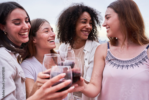 Four interracial female friends having fun together, toasting with red wine outdoors. Women enjoying on a rooftop celebrating and social gathering.