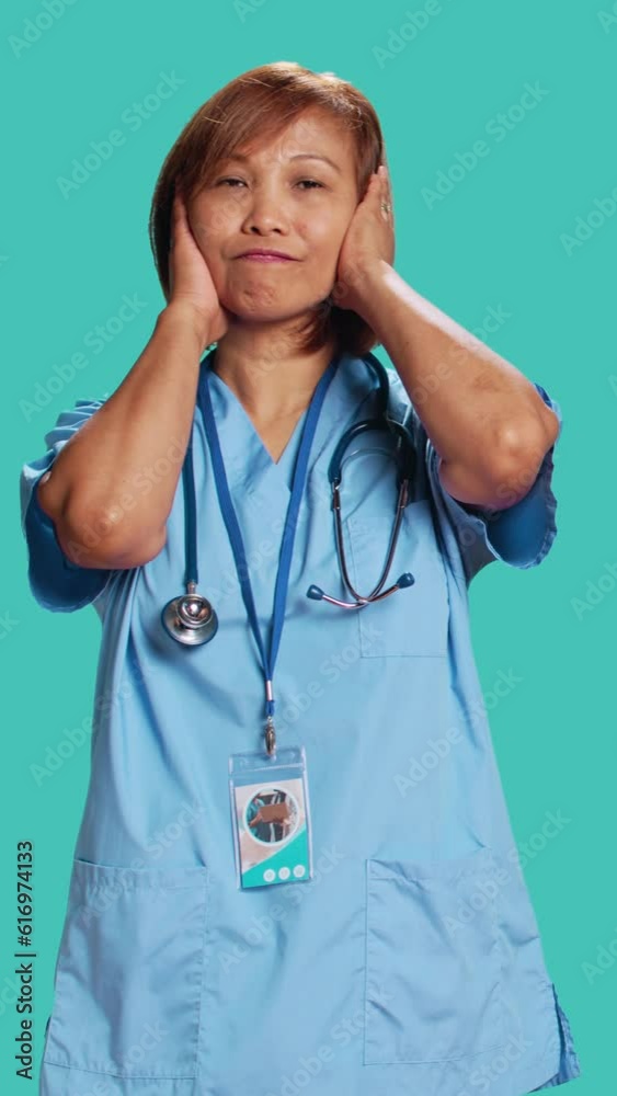 Certified nurse troubled by excessive noise while at work. Healthcare specialist covering ears with hands, annoyed by loud surrounding sounds, isolated over blue studio background Certified nurse troubled by excessive noise while at work. Healthcare specialist covering ears with hands, annoyed by loud surrounding sounds, isolated over blue studio background