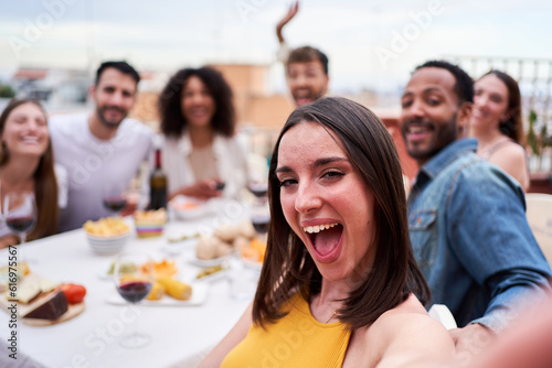 Group of multiethnic friends taking a selfie during social gathering in a rooftop. Cheerful photo of people dining outdoors in a terrace, looking at camera smiling.