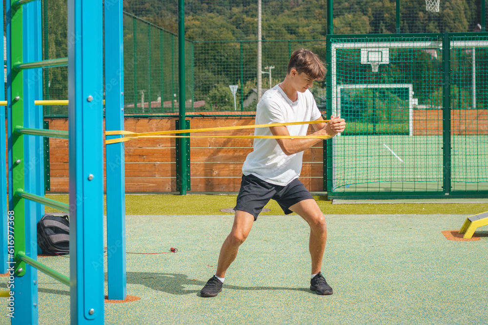 Obraz premium Goal-oriented teenager working on his body using resistance rubber performs strength training. Preparing for the sports season. Exercising with your own body. Outdoor workout playground. Biceps