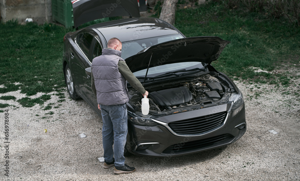 A man stands in front of a car hood.  Man fixing the car on the porch