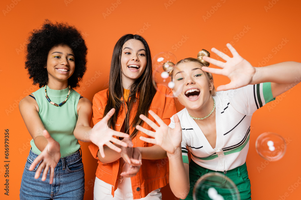Obraz premium Excited and cheerful multiethnic teen girls with bold makeup looking at soap bubbles while posing and standing on orange background, teen fashionistas with impeccable style concept
