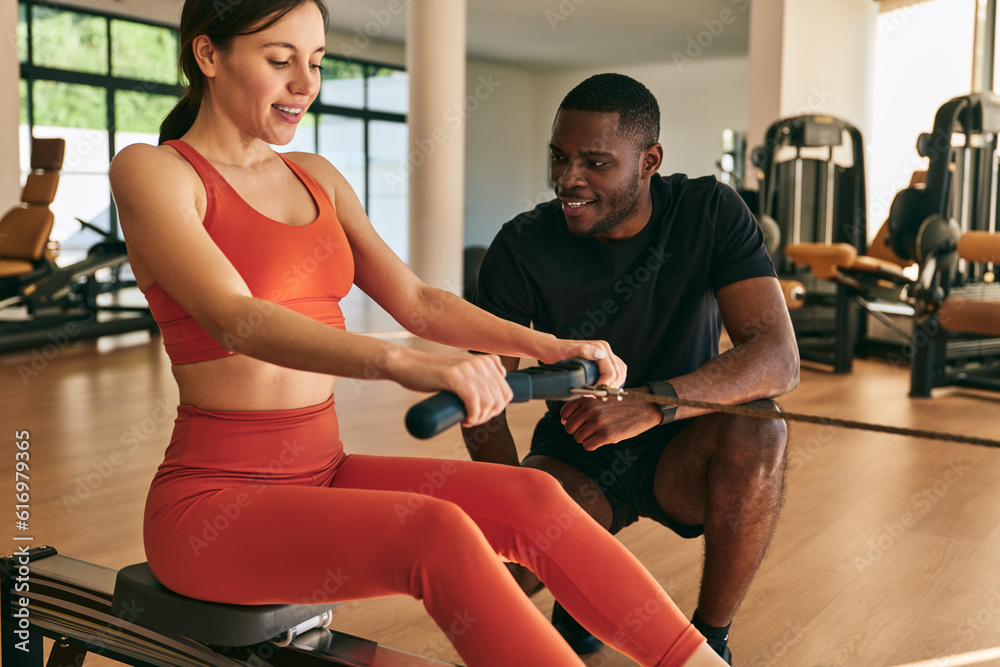 Cheerful sportswoman exercising on rowing machine with trainer Cheerful sportswoman exercising on rowing machine with trainer