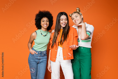 Cheerful and trendy multiethnic teenage girls with bold makeup and casual clothes posing with soap bubbles on orange background, teen fashionistas with impeccable style concept photo