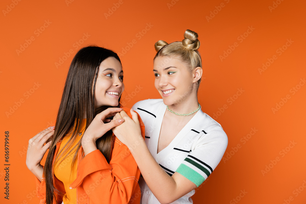 Smiling blonde teenage model with bold makeup hugging and looking at trendy brunette girlfriend while standing together isolated on orange, fashionable girls with sense of style Smiling blonde teenage model with bold makeup hugging and looking at trendy brunette girlfriend while standing together isolated on orange, fashionable girls with sense of style