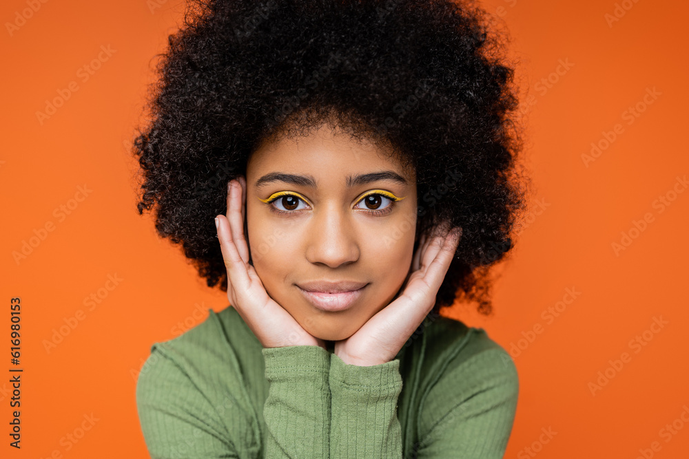 Portrait of teenage african american girl with bright makeup wearing green dress and looking at camera while touching cheeks isolated on orange, youth culture and generation z concept Portrait of teenage african american girl with bright makeup wearing green dress and looking at camera while touching cheeks isolated on orange, youth culture and generation z concept