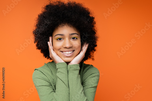 Portrait of happy teen african amerian girl with bold makeup wearing green dress and touching cheeks while standing isolated on orange, youth culture and generation z concept photo