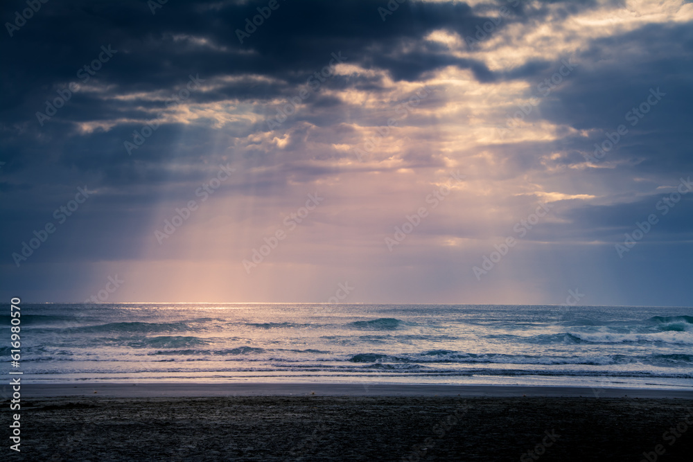 Sunset over Muriwai beach. Sun beams breaking through heavy clouds as waves splashing against the rocks. Auckland, New Zealand Sunset over Muriwai beach. Sun beams breaking through heavy clouds as waves splashing against the rocks. Auckland, New Zealand