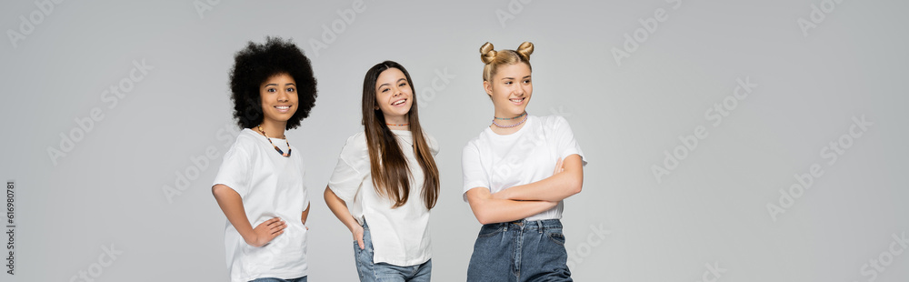 Joyful and multiethnic teenage girlfriends in white t-shirts posing and looking at camera while standing next to each other isolated on grey, lively teenage girls concept, banner Joyful and multiethnic teenage girlfriends in white t-shirts posing and looking at camera while standing next to each other isolated on grey, lively teenage girls concept, banner