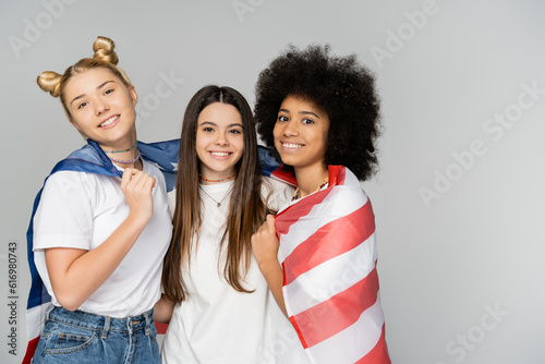Portrait of positive and multiethnic teenage girlfriends in white t-shirts covering with american flag and looking at camera isolated on grey, energetic teenage friends spending time