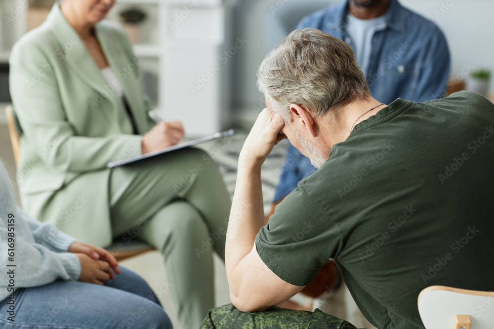 Focus on mature man puching his head while sitting in front of other people describing their problems to psychologist during session Focus on mature man puching his head while sitting in front of other people describing their problems to psychologist during session