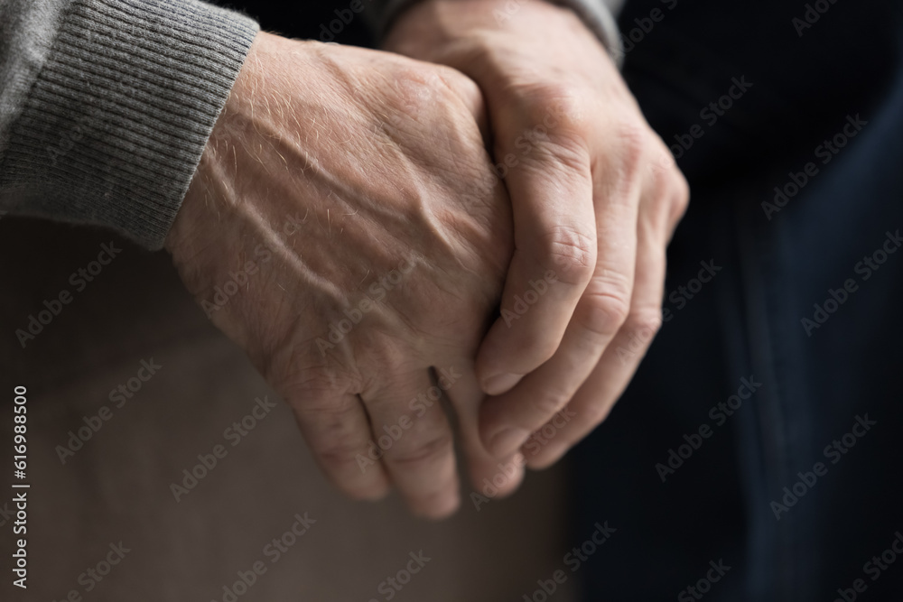 Fototapeta premium Close up upset old man sit on sofa alone with clenched hands, think, feels worried, anxious about future, spend life in nursing house. Elderly solitude, senile diseases and mental disorders of seniors