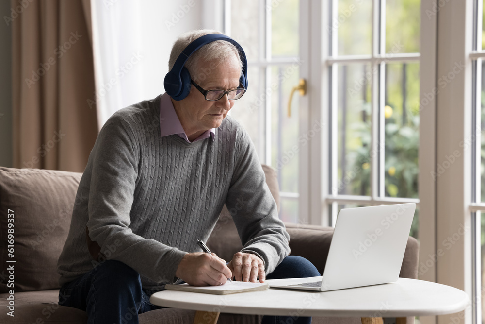 Serious older retired man in headphones e-learn, listen video lesson on laptop at home, take notes in copybook, watch webinar training, take part in virtual study, writes advices of online therapist Serious older retired man in headphones e-learn, listen video lesson on laptop at home, take notes in copybook, watch webinar training, take part in virtual study, writes advices of online therapist