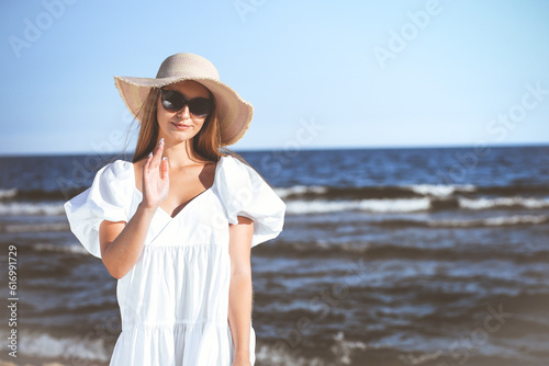 Happy blonde woman is posing on the ocean beach with sunglasses and a hat. Evening sun