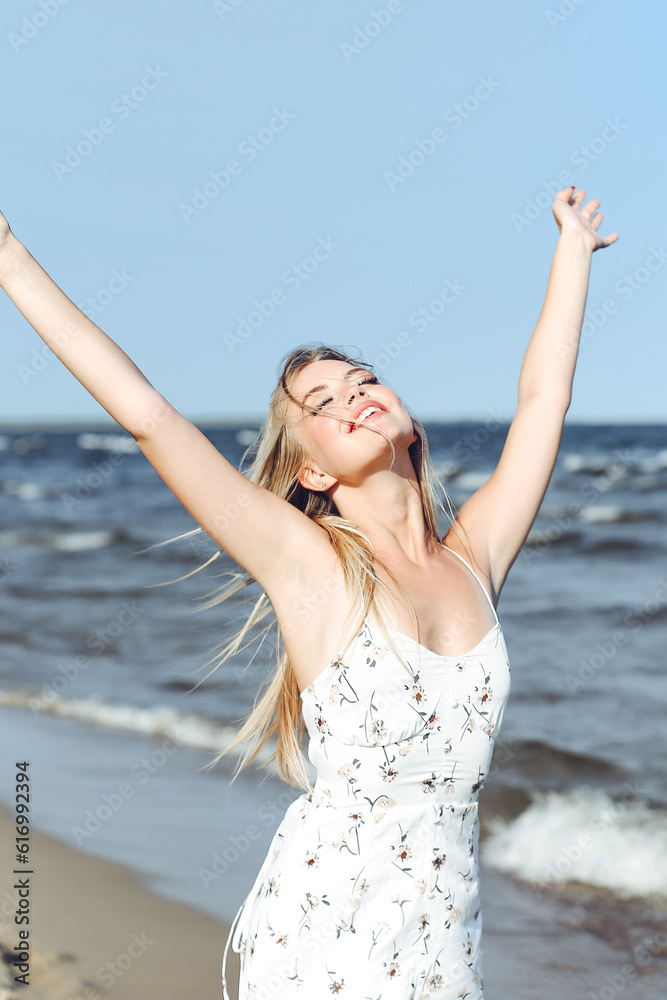 Happy blonde beautiful woman on the ocean beach standing in a white summer dress, raising hands Happy blonde beautiful woman on the ocean beach standing in a white summer dress, raising hands