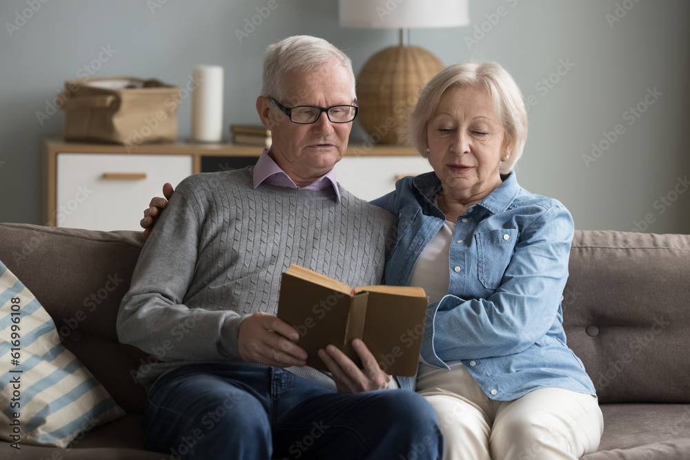 Focused mature spouses reading a book together at home. Serious baby-boomer generation spouses spend pastime resting on couch at home, enjoy favourite hobby, bestseller readers, literature store ad