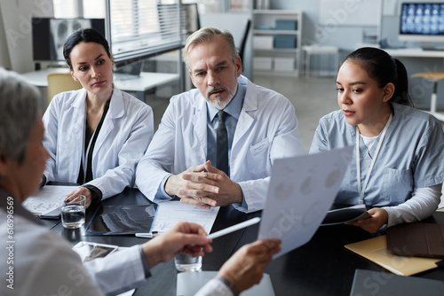 Focus on group of young and mature clinicians looking at medical data on document held by their colleague during report or presentation