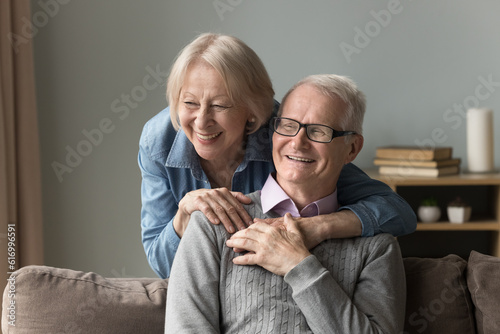 Laughing aged wife hugs his mature husband resting seated on sofa at home. Well-being and carefree life on retirement, enough pension for good and quality living, medical insurance cover ad for senior