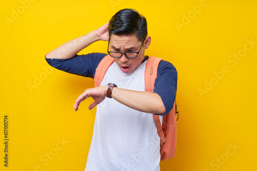Worried and shocked young Asian man student in casual clothes and glasses backpack scratching head and raising hand, looking watch isolated on yellow background. high school university college concept © Bangun Stock Photo