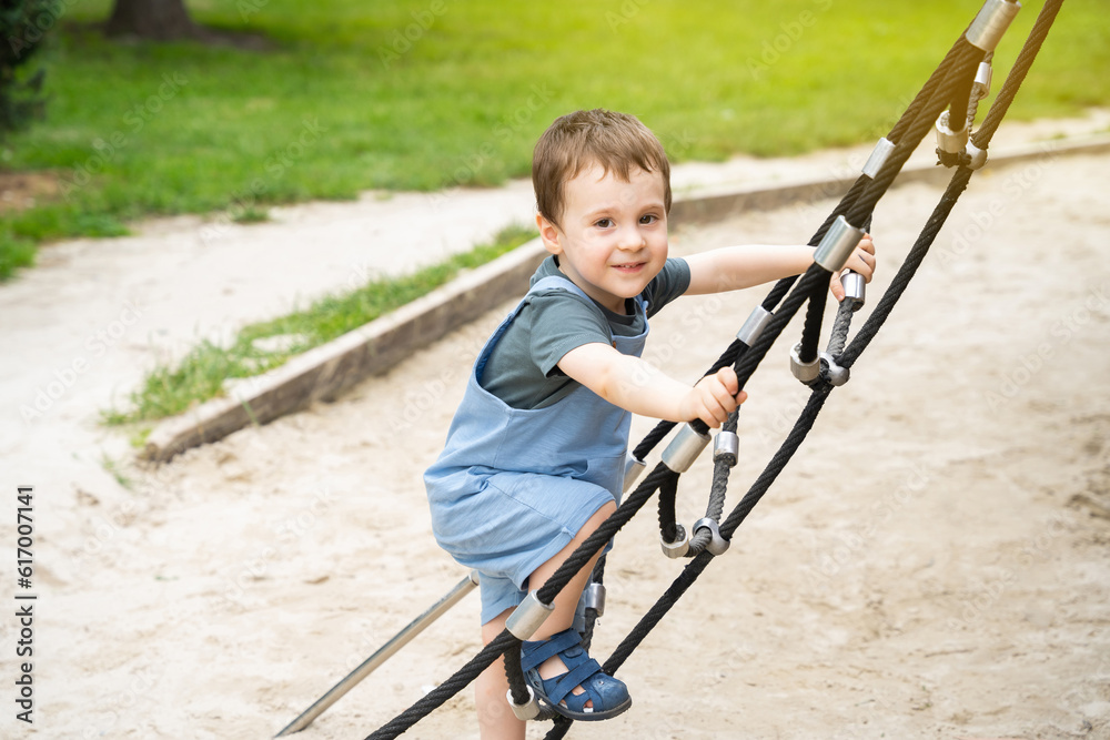 Happy toddler boy 2.5 years old climbs the rope ladder at the playground in the park. Active games and entertainment for children. sunny summer day Happy toddler boy 2.5 years old climbs the rope ladder at the playground in the park. Active games and entertainment for children. sunny summer day