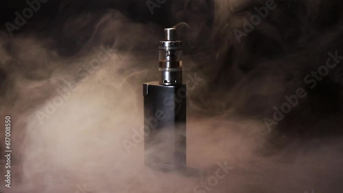 Vape is on a black table in clouds of thick heavy smoke on a black background with low light. Space for text.