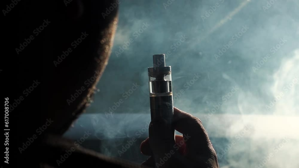 Young vaper man with beard vaping mechanical mod. Guy smokes an electronic cigarette by blowing a smoke vapor. Holds in hand Sitting at the wheel of a car. Space for text