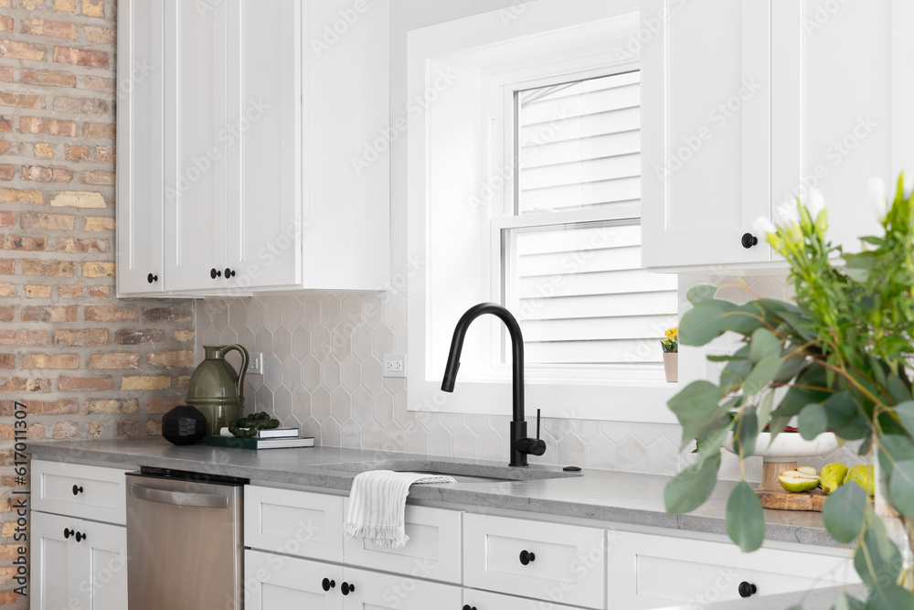 Fototapeta premium A beautiful kitchen sink detail with white cabinets, marble countertops, and a hexagon tiled backsplash. A blurred plant in the foreground.