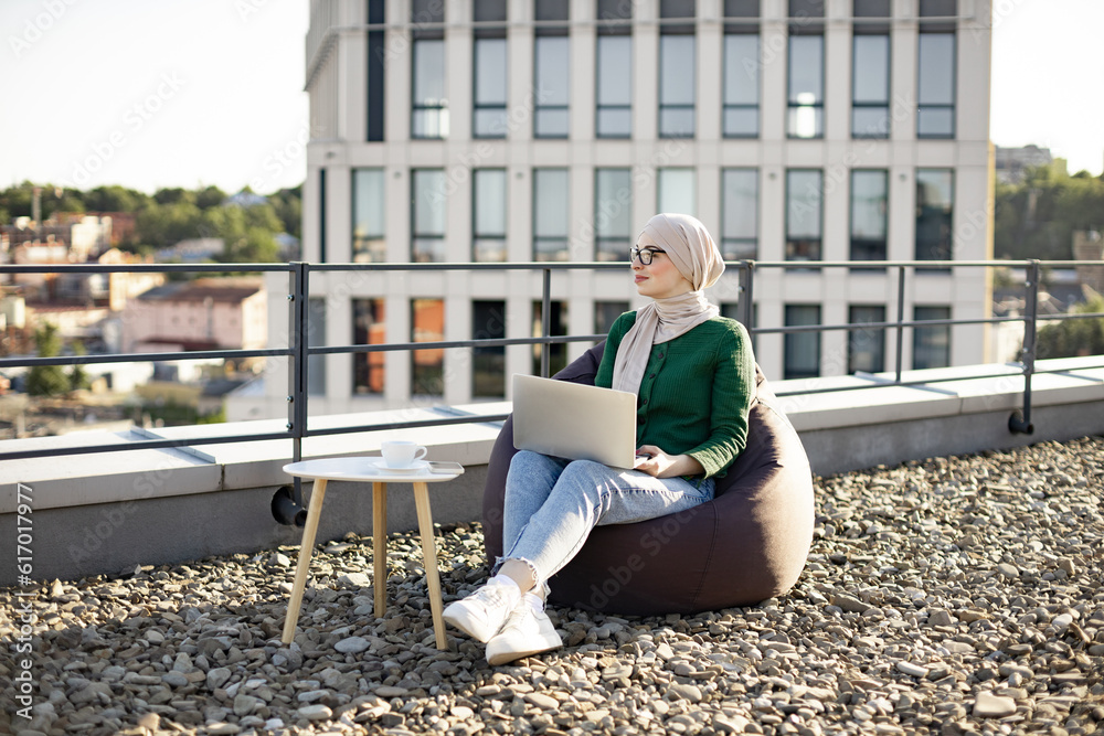 Beautiful woman wearing glasses and hijab daydreaming while holding laptop on knees in pouf chair outdoors. Arabian freelancer analyzing new project while staying on office roof terrace on sunny day. Beautiful woman wearing glasses and hijab daydreaming while holding laptop on knees in pouf chair outdoors. Arabian freelancer analyzing new project while staying on office roof terrace on sunny day.