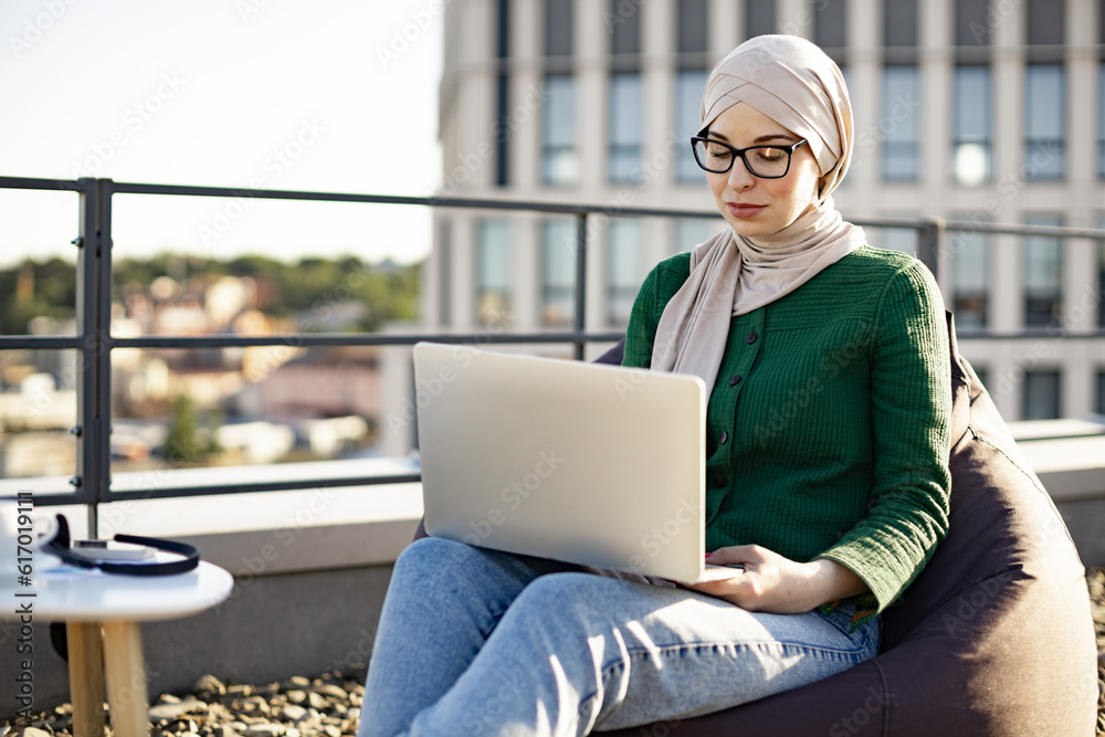 Smiling arabian lady in hijab sitting in beanbag chair near coffee table with drink and devices in fresh air outdoors. Cheerful businesswoman in glasses starting working day with laptop on terrace. Smiling arabian lady in hijab sitting in beanbag chair near coffee table with drink and devices in fresh air outdoors. Cheerful businesswoman in glasses starting working day with laptop on terrace.