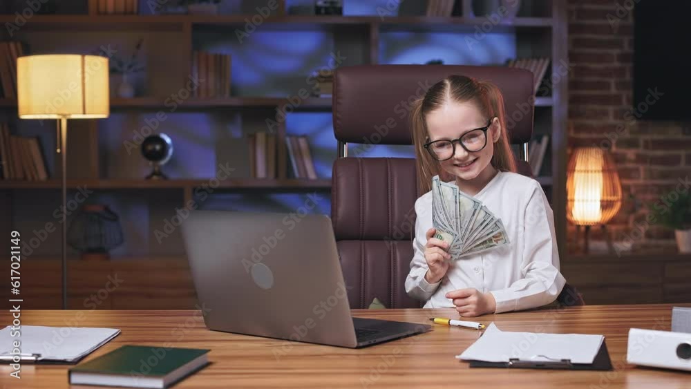 Cute little school girl playing with dollar banknotes while sitting at office table with personal computer in comfy workplace. Pretty kid wearing eyeglasses, having fun and covering face with money. Cute little school girl playing with dollar banknotes while sitting at office table with personal computer in comfy workplace. Pretty kid wearing eyeglasses, having fun and covering face with money.