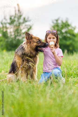 Cute little girl eating ice cream and hugging a shepherd dog sitting on the grass in the park in summer. High quality photo, blurred background, vertical orientation © Serhii