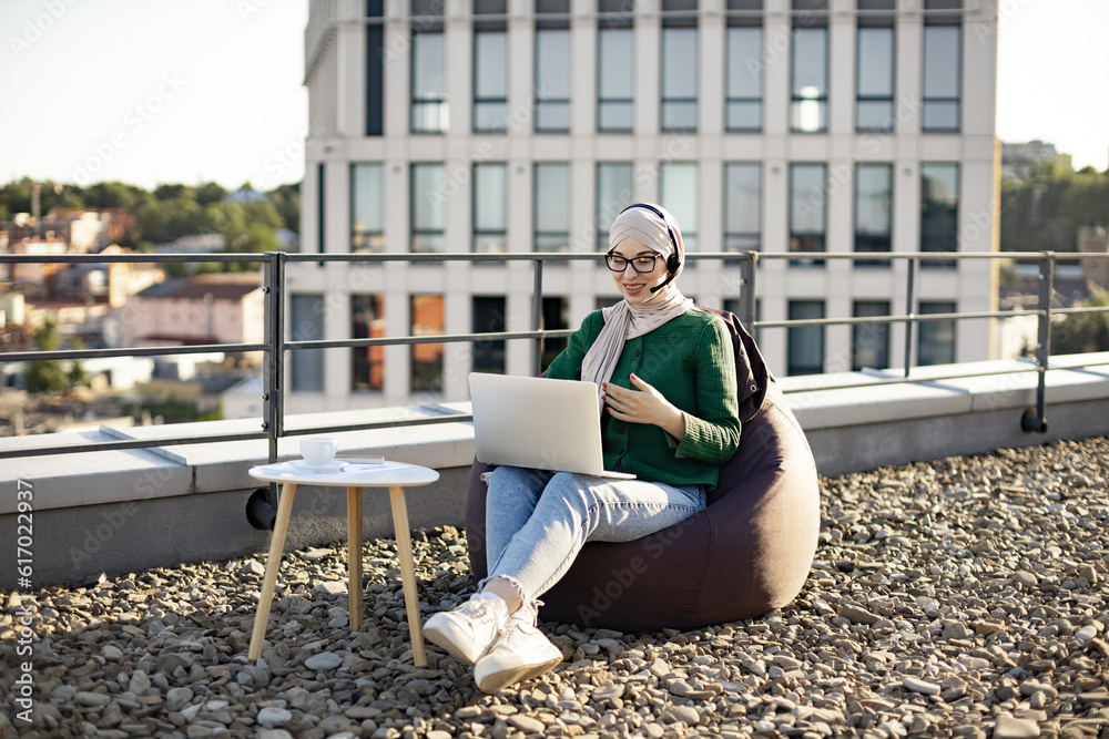 Relaxed muslim woman in headscarf conducting web conference via laptop and headset while staying on flat roof. Professional entrepreneur working outside of corporate offices via wireless devices. Relaxed muslim woman in headscarf conducting web conference via laptop and headset while staying on flat roof. Professional entrepreneur working outside of corporate offices via wireless devices.