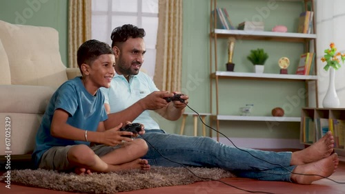 Happy excited indian father with son playing video game using joystick on floor at home - concept of Joyful playtime, weekdn fun and relationship bonding photo