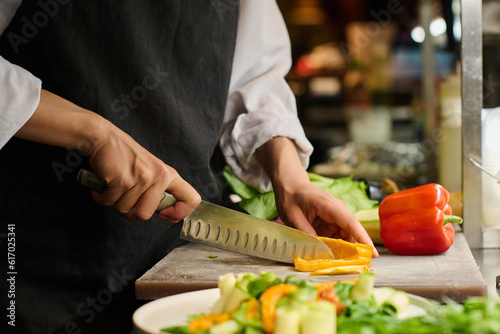 Close-up of hands of young female kitchen worker chopping fresh yellow pepper for salad or some other dish while standing by workplace