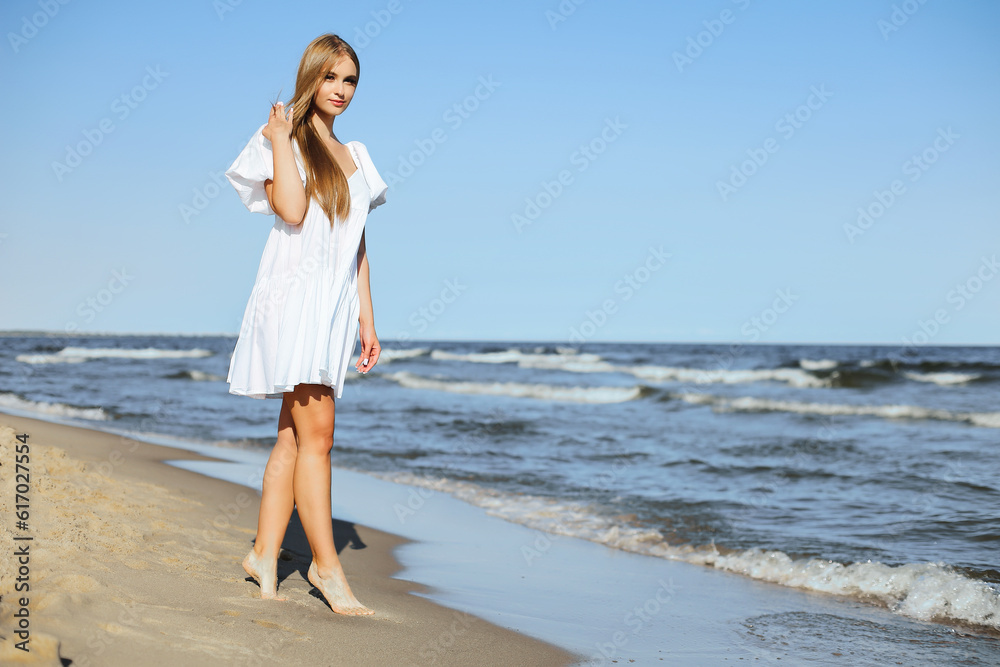 Happy smiling beautiful woman is walking on the ocean beach in a white summer dress