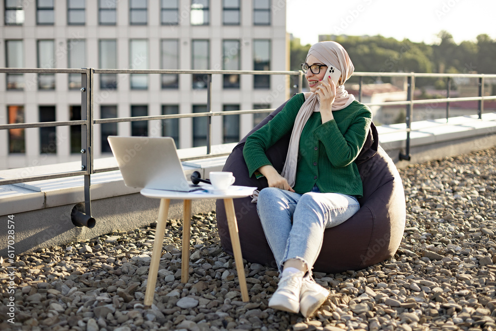 Carefree arabian lady in casual wear and hijab making phone call while relaxing in beanbag on flat roof. Beautiful adult female in spectacles discussing agreement while doing outdoor office work.