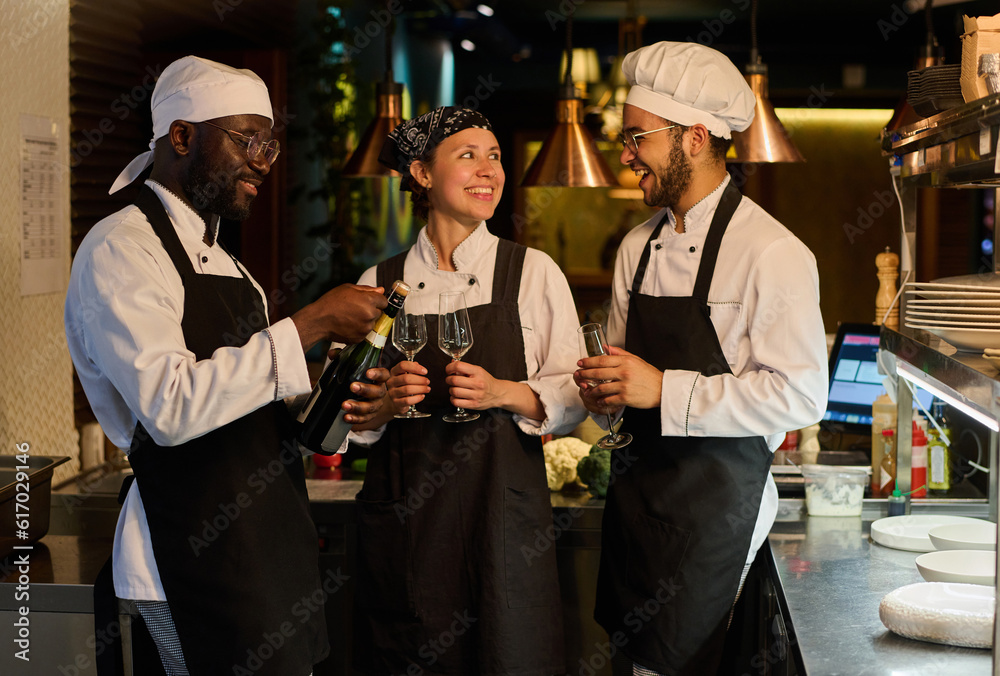 Group of happy young intercultural kitchen workers in uniform celebrating start of new cafe while one of them opening bottle of champagne