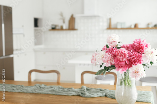 Beautiful peonies in vase on wooden table on background of stylish white kitchen with appliances in new scandinavian house. Modern kitchen interior and summer floral arrangement