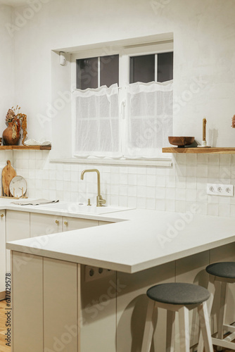 Modern kitchen interior. Stylish white kitchen cabinets, brass faucet and granite sink, wooden shelves with utensils and evening light in new scandinavian house. Modern minimal kitchen design