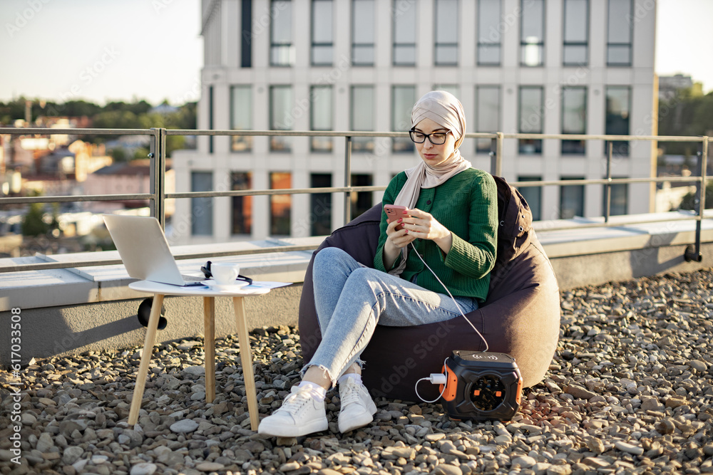 Focused arabian lady in hijab holding cell phone connected to portable power station via charging cord on roof terrace. Efficient freelancer working remotely using eco-friendly energy source outside.