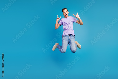 Full length photo of excited young man in t-shirt jumping celebrating party horned symbol isolated blue background